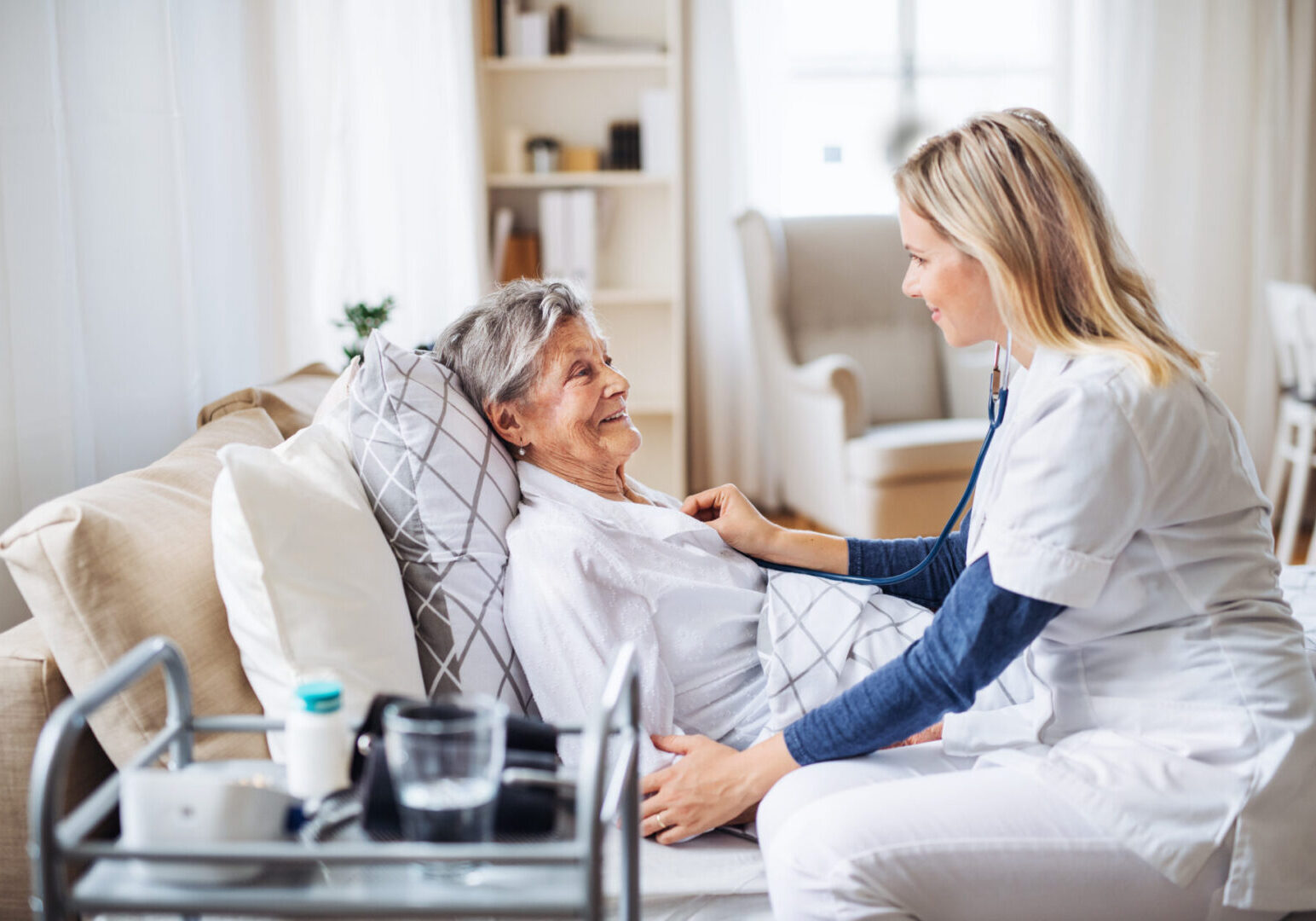 A health visitor examining a sick senior woman lying in bed at home with stethoscope