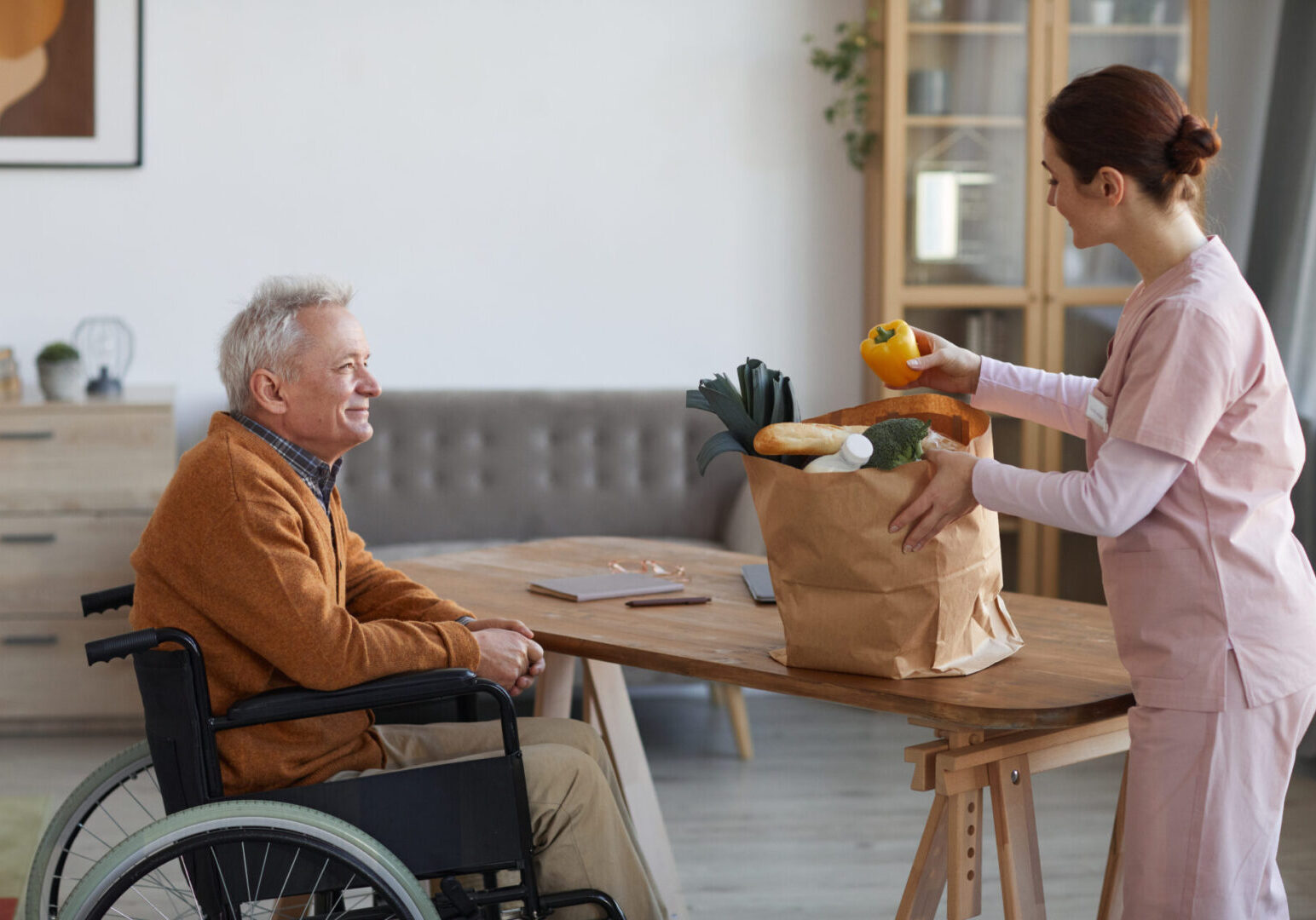 Side view portrait of smiling senior man in wheelchair looking at female nurse bringing groceries