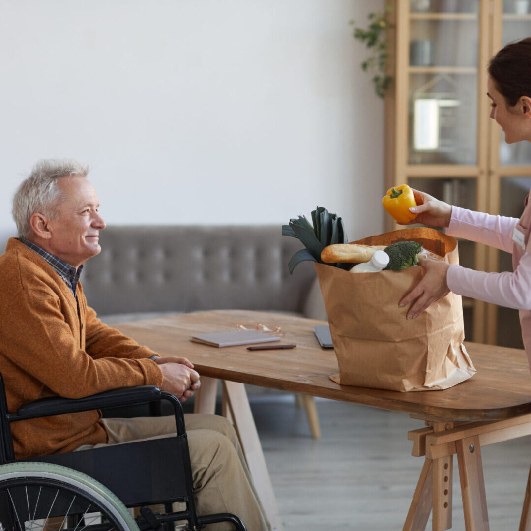 Side view portrait of smiling senior man in wheelchair looking at female nurse bringing groceries